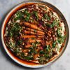 An aerial view of a plate of honey roasted carrots atop a FODMAP friendly tofu hummus. The hummus is dotted with smoky roasted chickpeas, green tahini sauce, chopped almonds and rose petals. The dish is surrounded by a sea of bright red chilli oil. It sits atop a white ceramic plate on a white marble table.