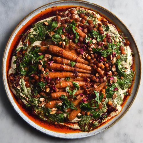 An aerial view of a plate of honey roasted carrots atop a FODMAP friendly tofu hummus. The hummus is dotted with smoky roasted chickpeas, green tahini sauce, chopped almonds and rose petals. The dish is surrounded by a sea of bright red chilli oil. It sits atop a white ceramic plate on a white marble table.