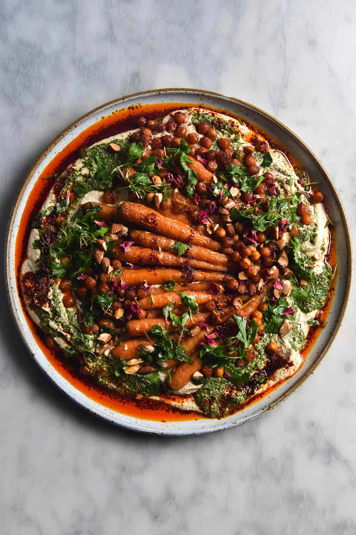 An aerial view of a plate of honey roasted carrots atop a FODMAP friendly tofu hummus. The hummus is dotted with smoky roasted chickpeas, green tahini sauce, chopped almonds and rose petals. The dish is surrounded by a sea of bright red chilli oil. It sits atop a white ceramic plate on a white marble table.