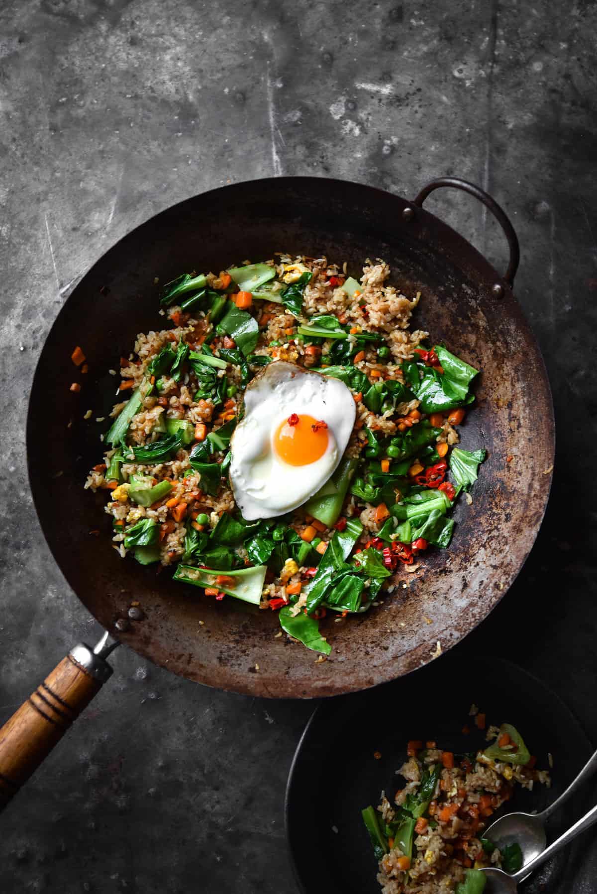An aerial image of a black wok filled with low FODMAP fried rice on a dark blue steel backdrop. A plate of fried rice sits to the bottom right of the image and the fried rice is topped with a fried egg.