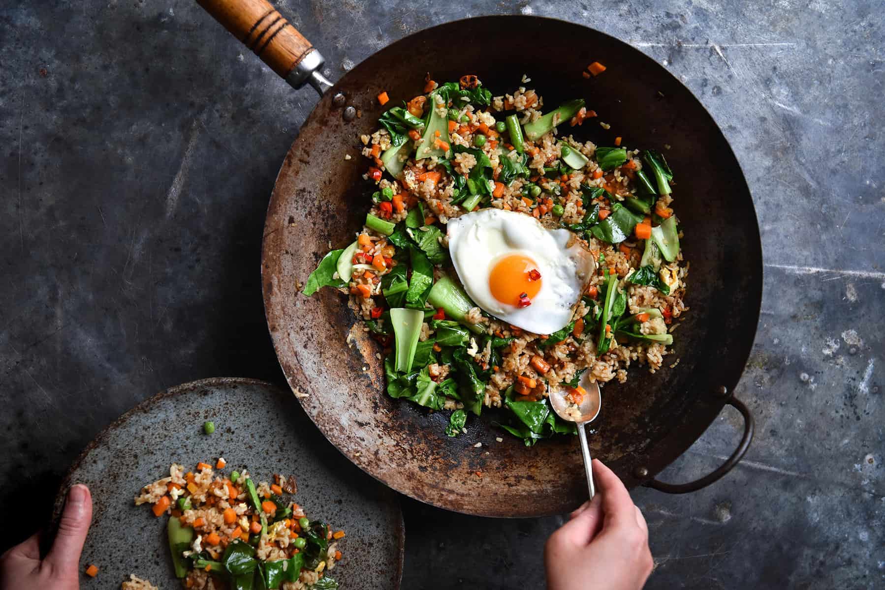 An aerial image of a dark rusty wok filled with low FODMAP fried rice atop a dark blue steel backdrop. A plate of fried rice sits to the left of the of wok and a hand extends out scoop more fried rice onto the plate. 