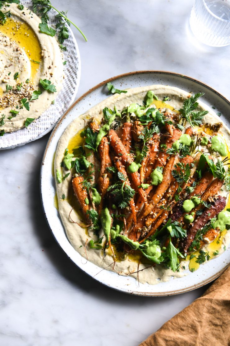 An aerial view of a dukkah and honey roasted carrot salad sitting on a bed of hummus and topped with a green tahini sauce. The main salad sits on a white ceramic plate on a white marble table, and a second small plate of hummus sits to the top left of the image.