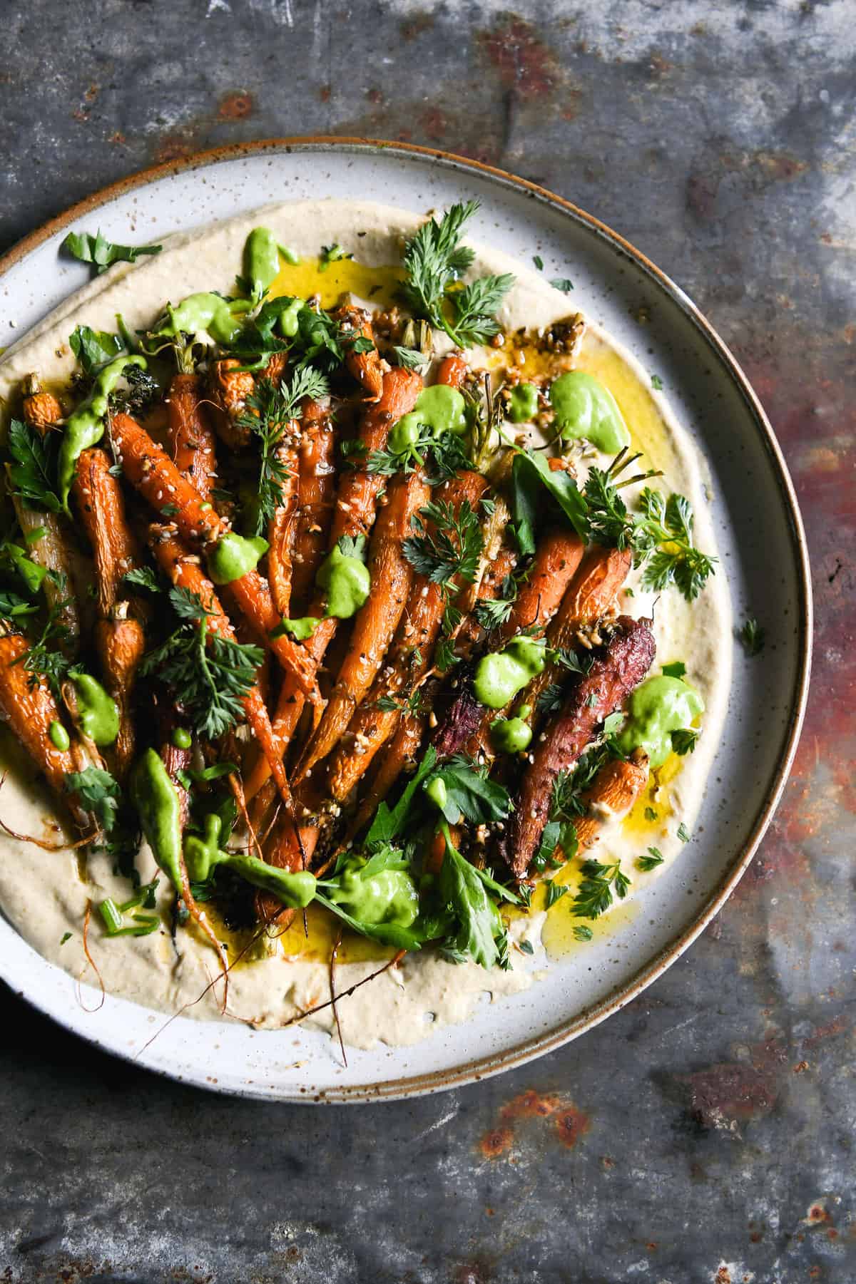 Dukkah and honey roasted carrot salad with FODMAP friendly hummus and a green herb sauce on a white ceramic plate atop a dark blue steel backdrop