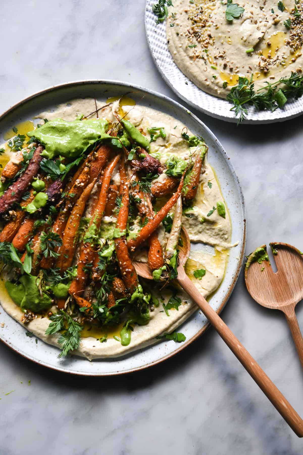 An aerial view of a dukkah and honey roasted carrot salad sitting on a bed of hummus and topped with a green tahini sauce. The main salad sits on a white ceramic plate on a white marble table, and a second small plate of hummus sits to the top right of the image.