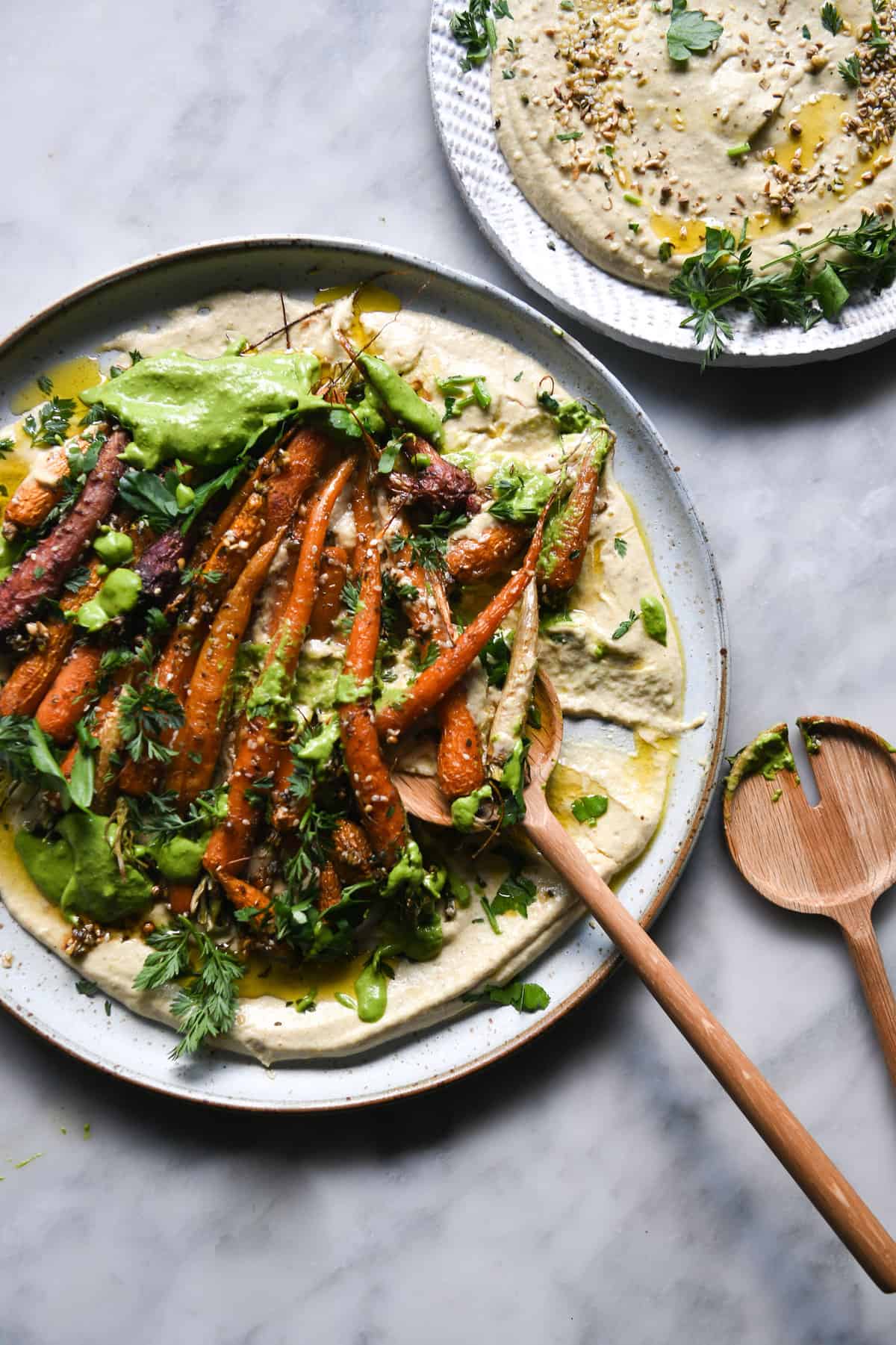 An aerial image of a dukkah and honey roasted carrot salad with FODMAP friendly hummus and a green herb sauce on a white ceramic plate atop a white marble table. 