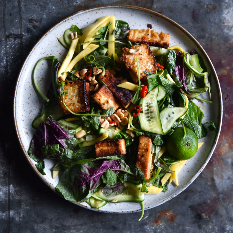 An aerial image of a fresh summer salad with crispy oven baked tofu. The salad sits on a white ceramic plate atop a dark blue mottled steel backdrop.