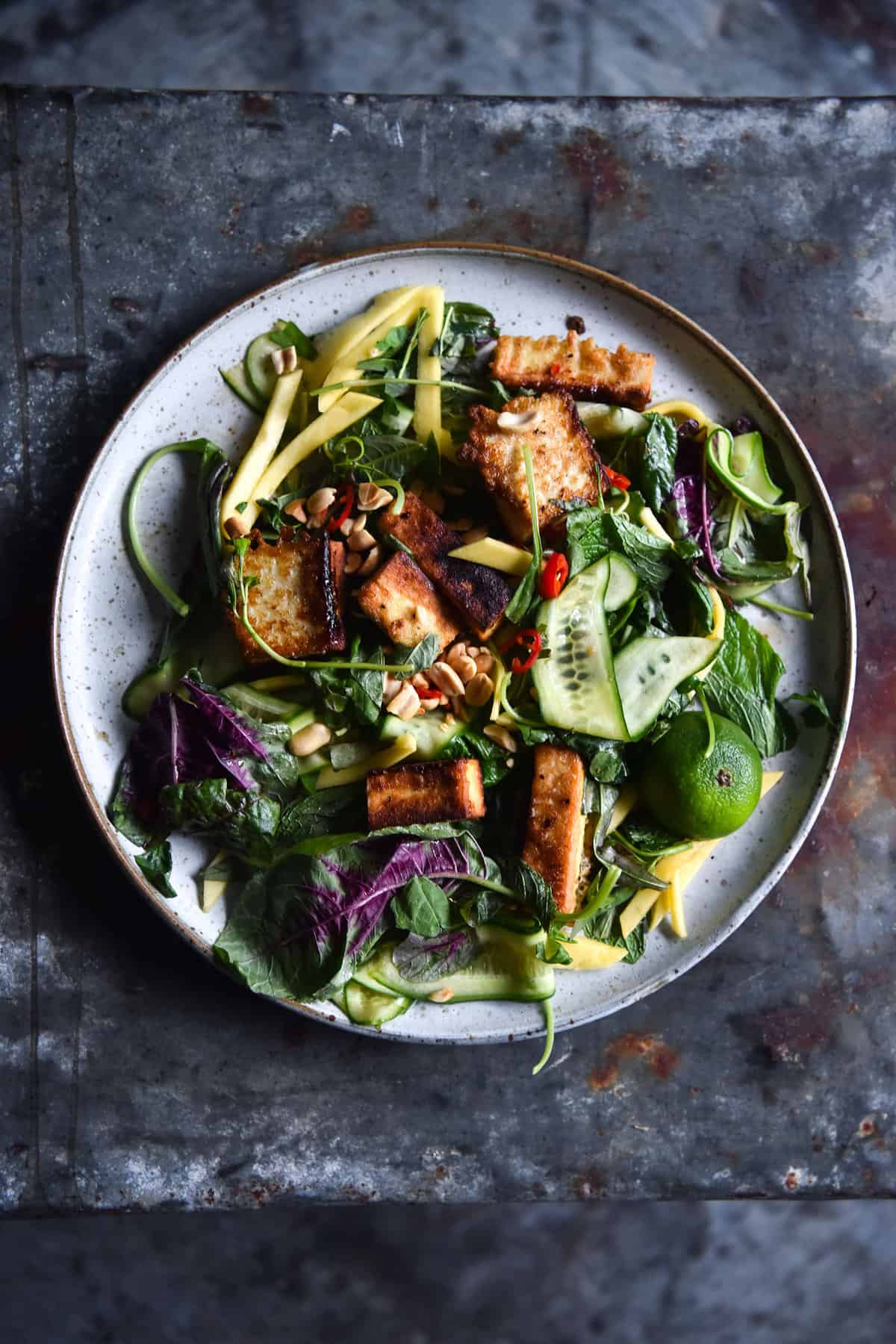 An aerial image of a fresh summer salad with crispy oven baked tofu. The salad sits on a white ceramic plate atop a dark blue mottled steel backdrop.