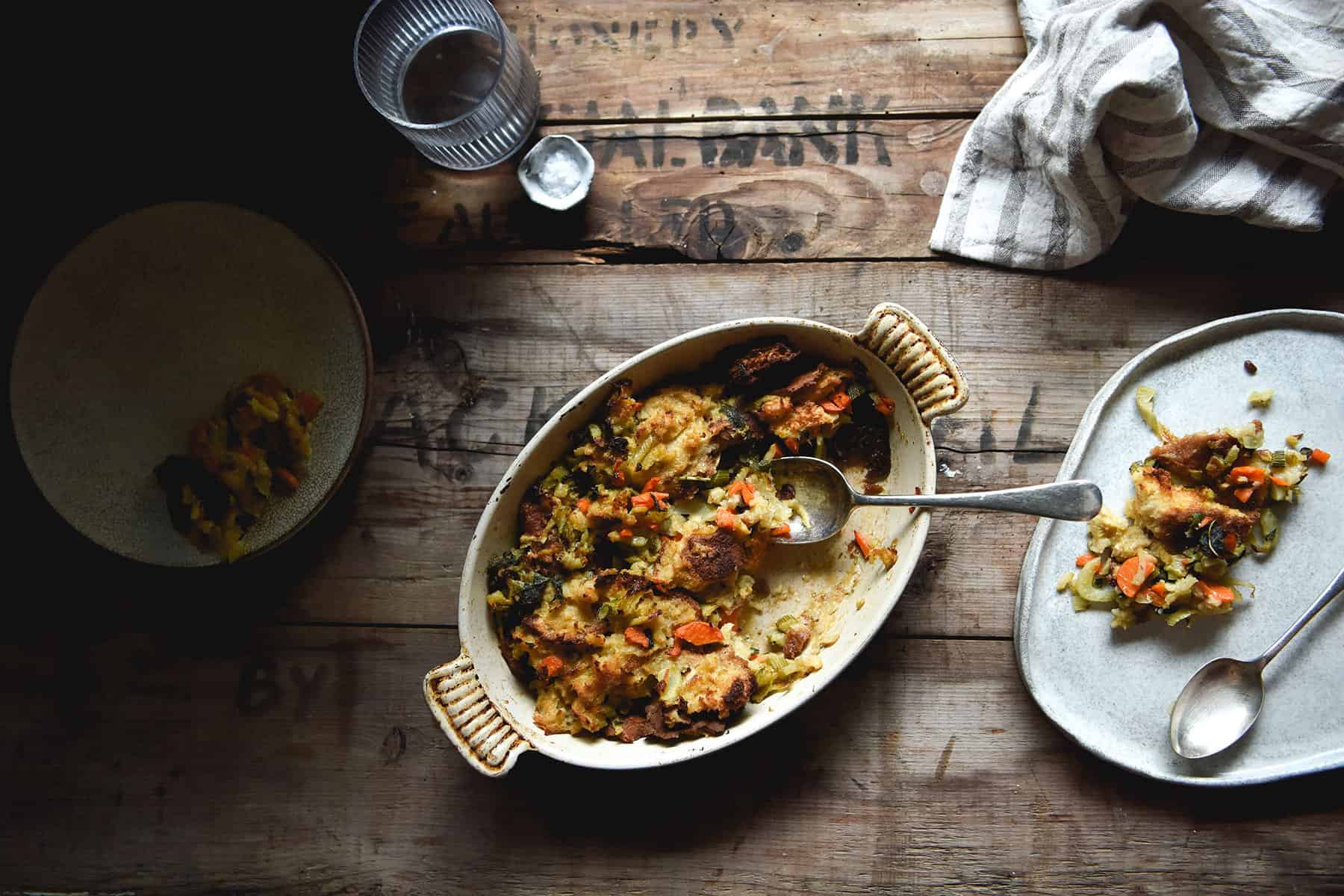 An aerial image of FODMAP friendly, gluten free vegan stuffing in a scalloped baking dish atop a moody wooden table. The dish of stuffing is surrounded by extra plates and neutral toned linen tea towels.