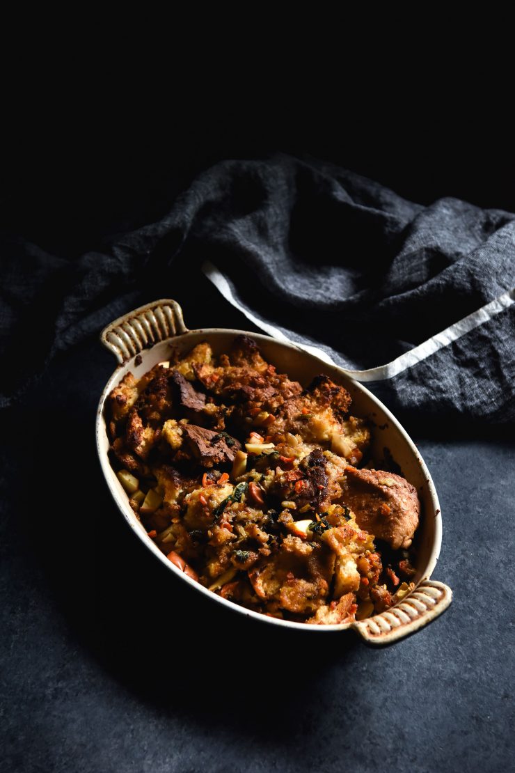 A moody photo of a baking dish filled with gluten free stuffing. The dish sits atop a dark blue backdrop and a blue linen cloth sits in the background