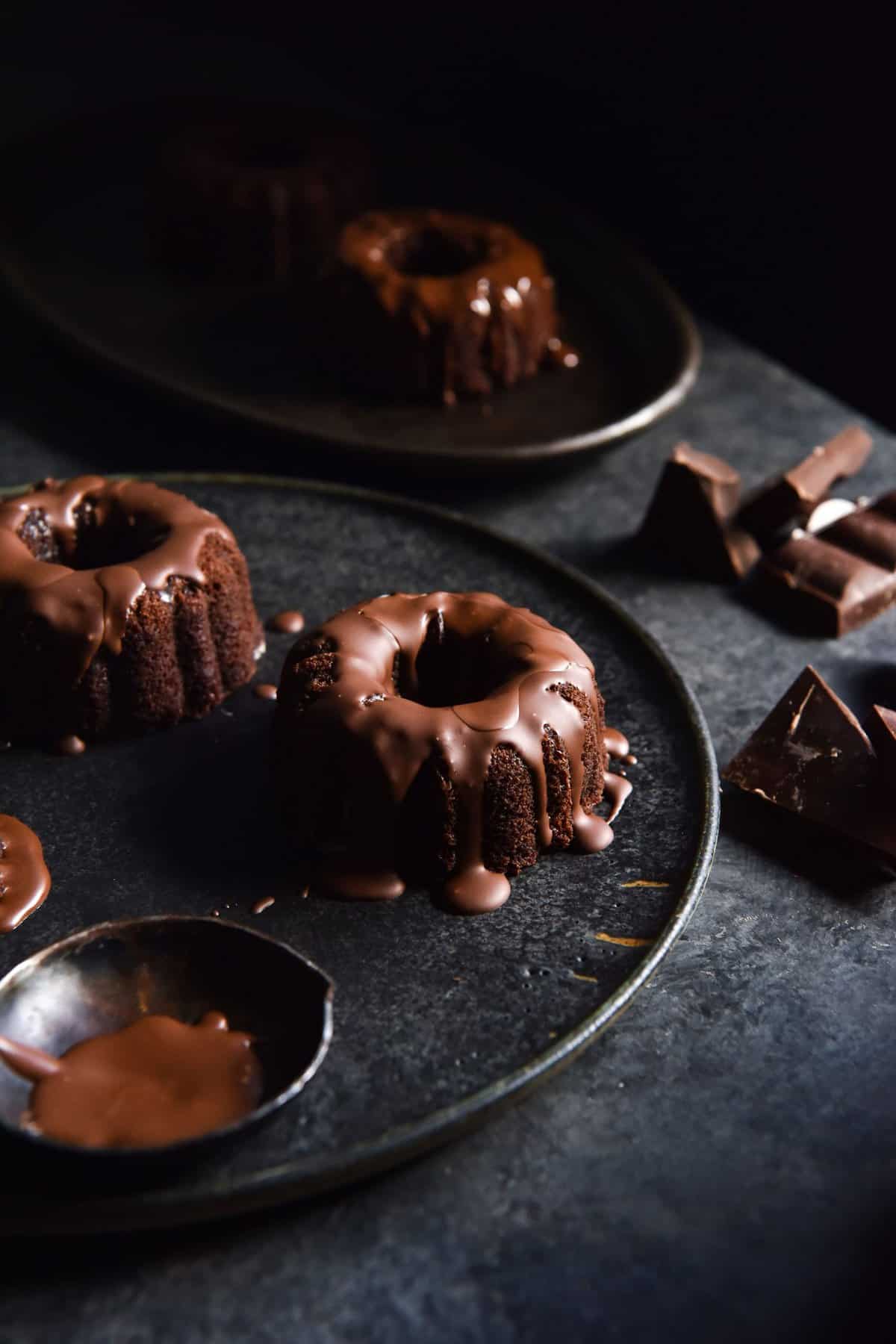 A moody side on image of flourless chocolate bundt cakes on a dark grey plate atop a dark blue backdrop. The bundt cakes are topped with extra melted chocolate.