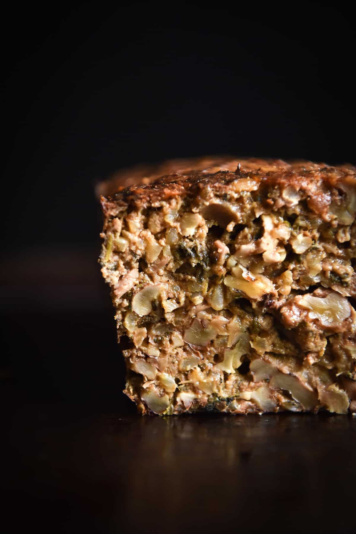 A side on image of a FODMAP friendly gluten free nut loaf against a black backdrop. The first slice has been taken, revealing the soft and meaty loaf texture. 