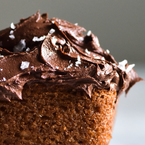 A close up, side on image of a grain free vanilla coconut cake topped with chocolate buttercream and sea salt flakes. The cake sits on a white marble table against a white backdrop.