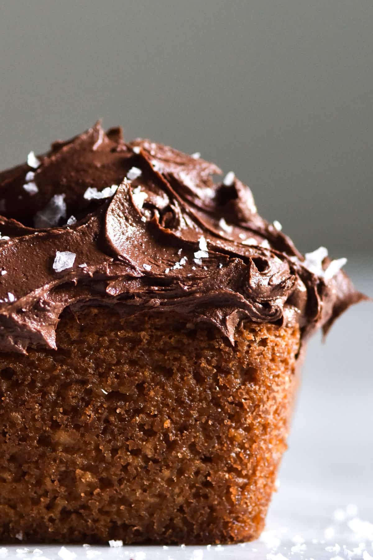 A close up, side on image of a grain free vanilla coconut cake topped with chocolate buttercream and sea salt flakes. The cake sits on a white marble table against a white backdrop.