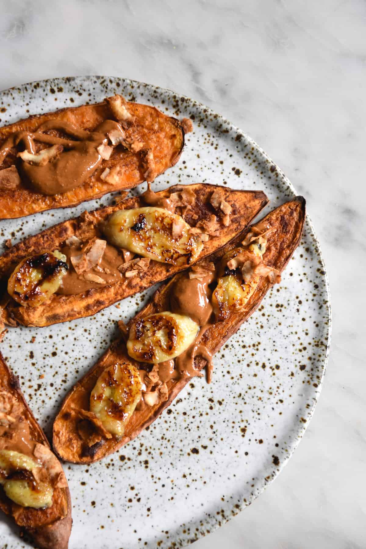 An aerial image of sweet potato 'toast' topped with almond butter and banana on a white ceramic plate atop a white marble table.