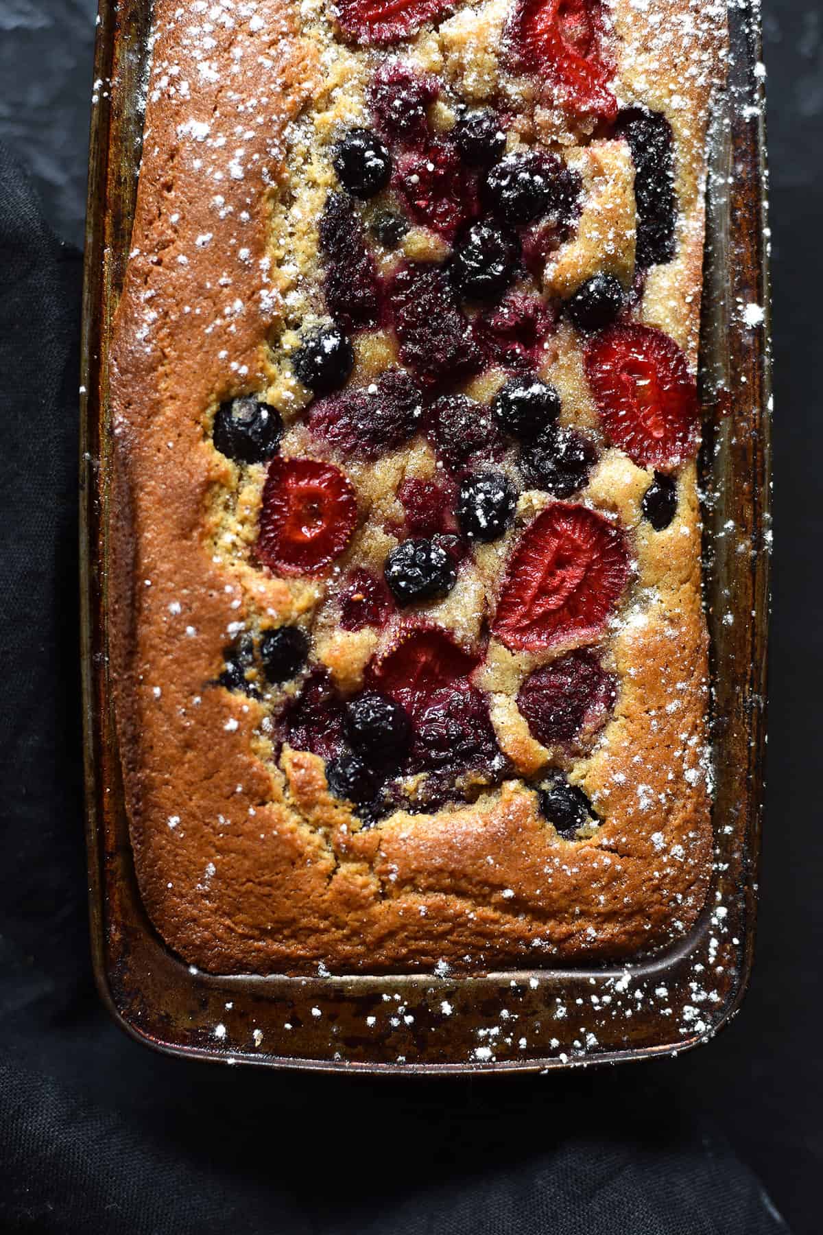 An aerial, close up view of a grain free vanilla coconut cake topped with mixed berries. The loaf sits in a baking tin on a dark backdrop