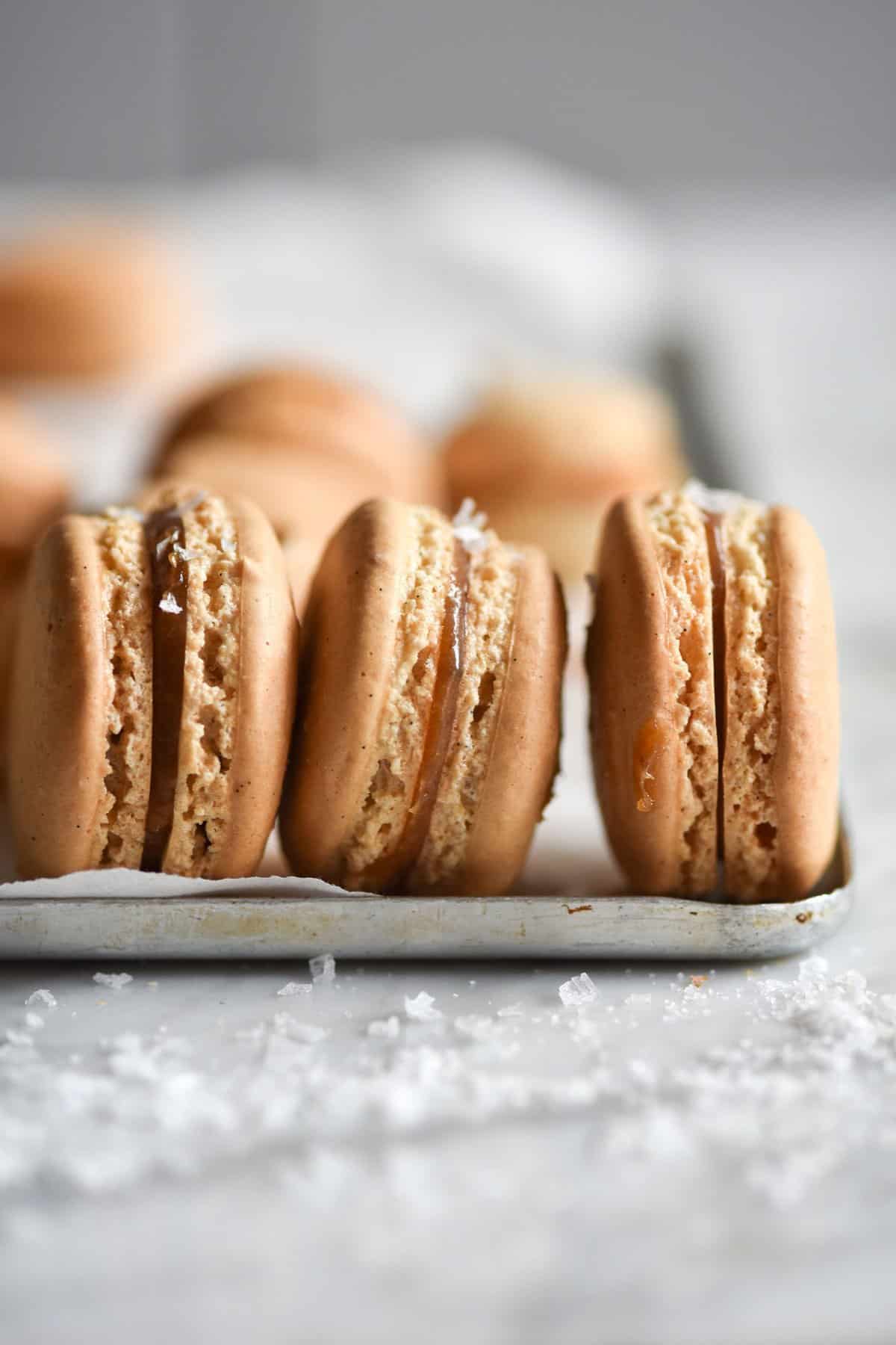 A side on macro image of banana and salted caramel macarons on a lined baking sheet atop a white marble table.