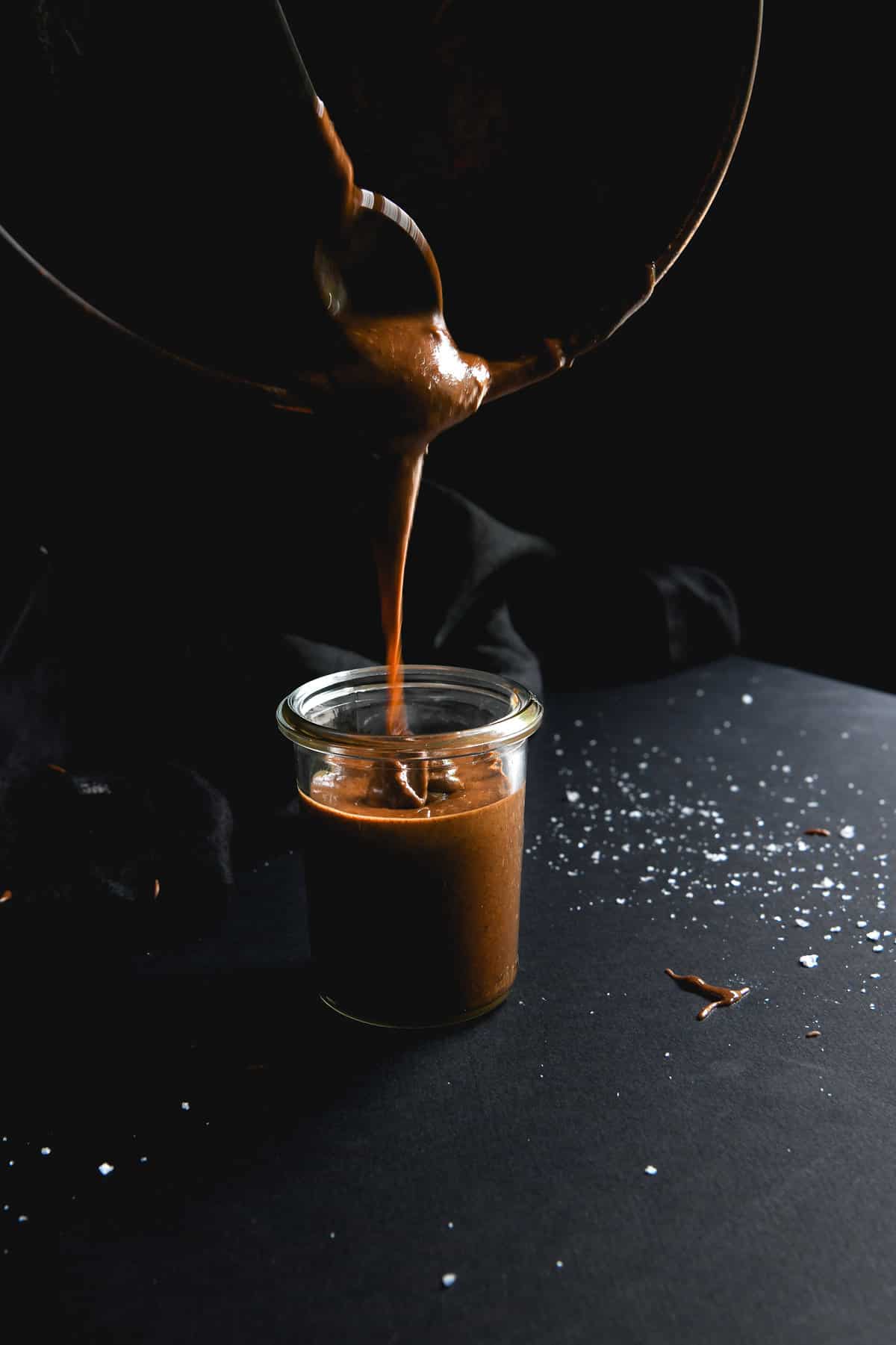 A moody side on image of dark roasted almond butter being poured into a glass container.