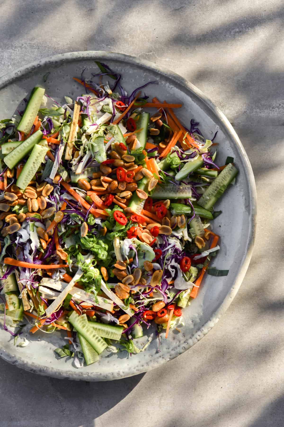 An aerial view of a Vegan Vietnamese coleslaw salad, topped with lots of peanuts, chilli and spring onion greens. The serving platter is on grey tiles in dappled sunlight