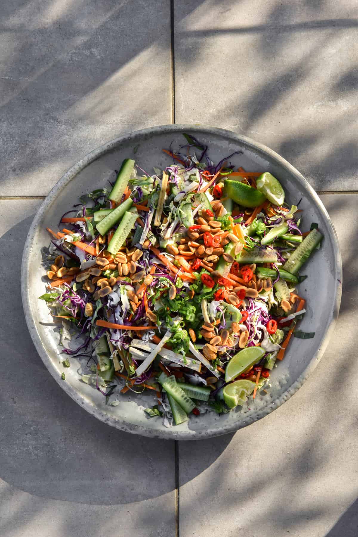 An aerial view of a Vegan Vietnamese coleslaw salad, topped with lots of peanuts, chilli and spring onion greens. The serving platter is on grey tiles in dappled sunlight
