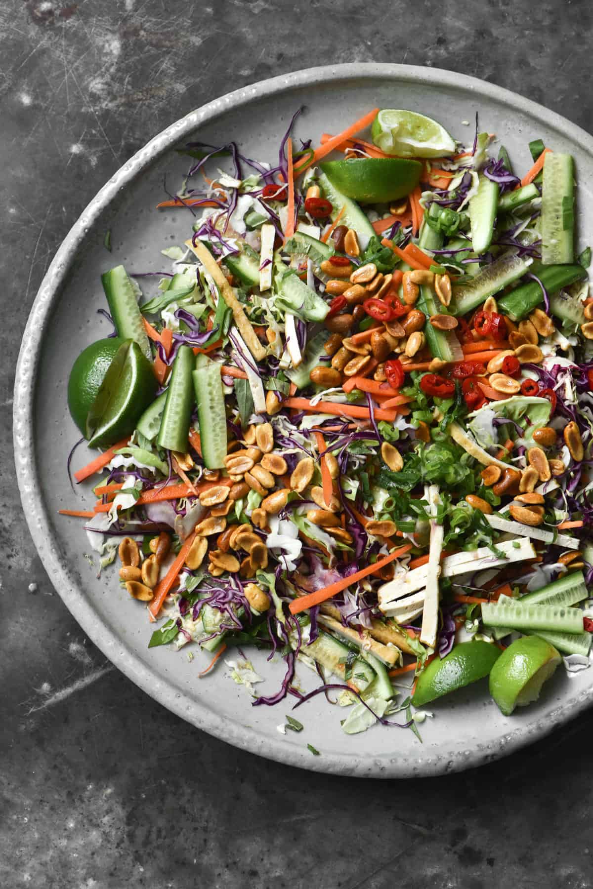 An aerial view of a big ceramic plate of colourful Vegan Vietnamese coleslaw. The coleslaw is topped with lots of lime wedges, toasted peanuts and spring onion greens. The coleslaw is set against a mottled blue steel backdrop