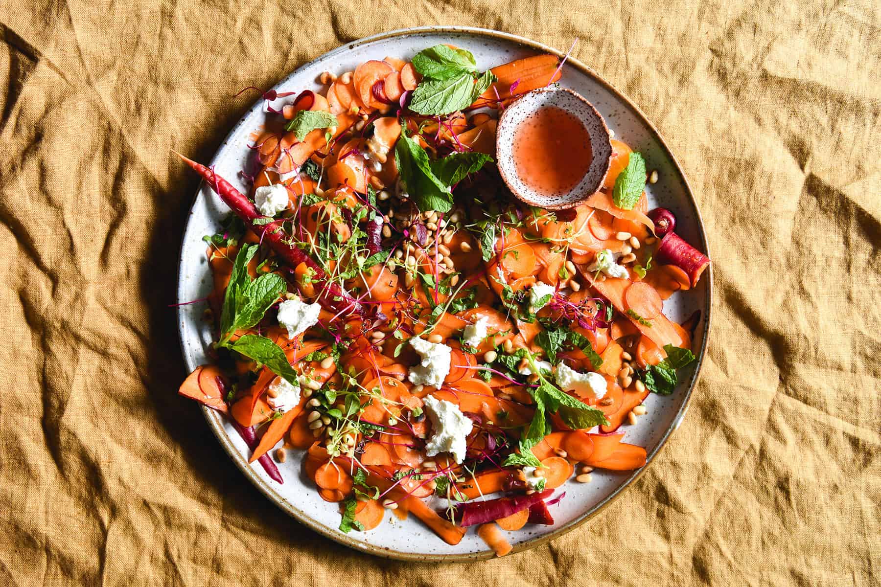 Shaved carrot salad with goat's cheese, toasted pine nuts, mint and an orange blossom, lemon and Campari dressing on a white ceramic plate atop a turmeric coloured linen tablecloth.