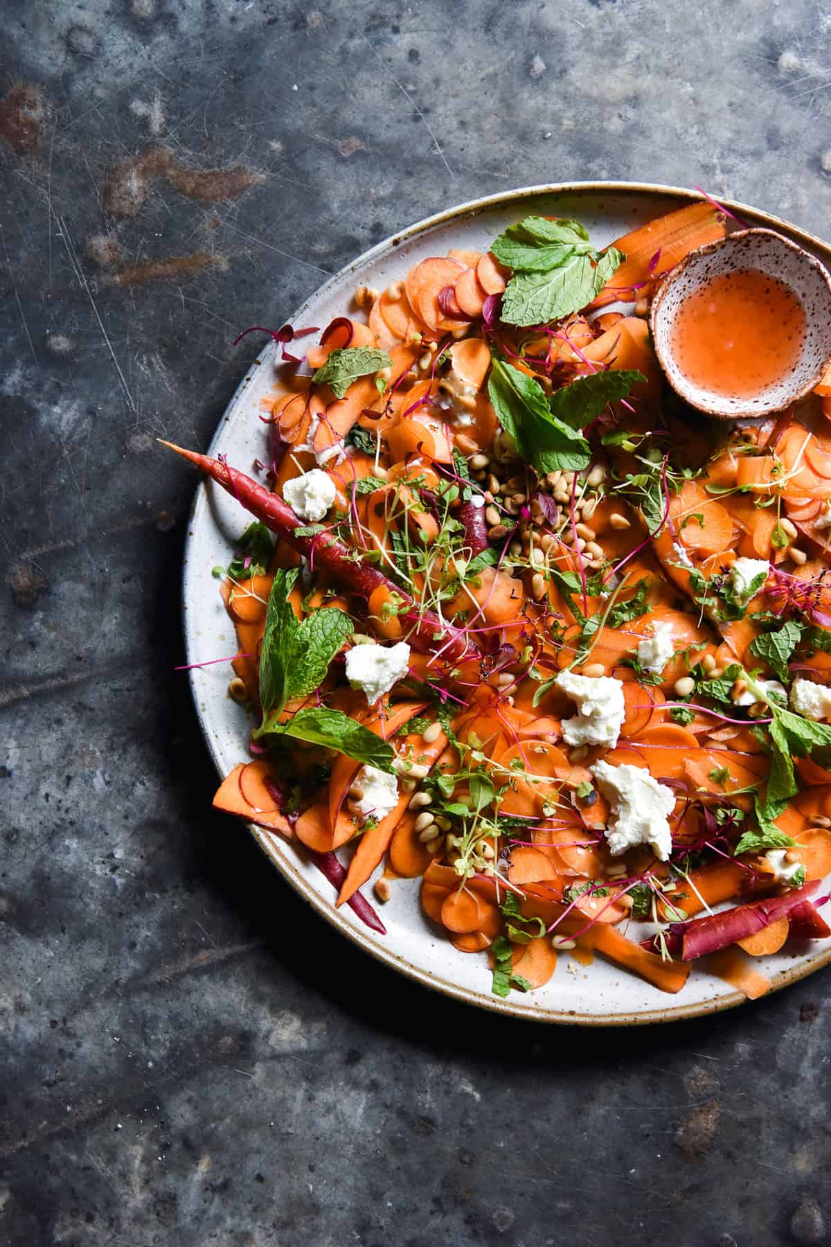 An aerial image of a shaved carrot salad on a white ceramic plate atop a dark blue steel backdrop. The salad is topped with mint, microherbs, and pine nuts and a small white ceramic bowl of dressing sits to the top right.