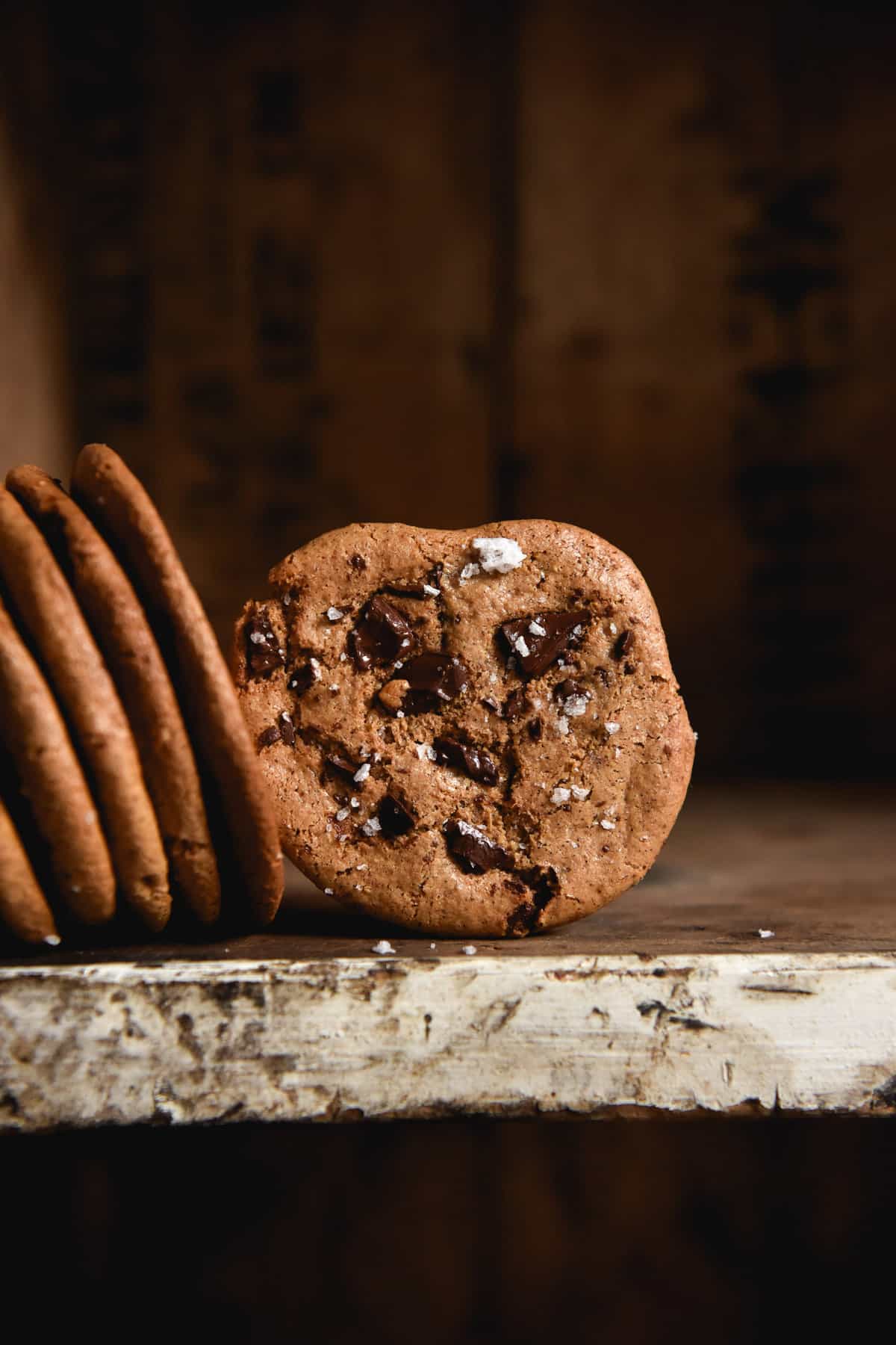 A moody side on image of an almond butter cookie on a dark coloured shelf. The central cookie faces the camera and is studded with chocolate chips. The remaining cookies lean up against the shelf to the left of the central cookie.