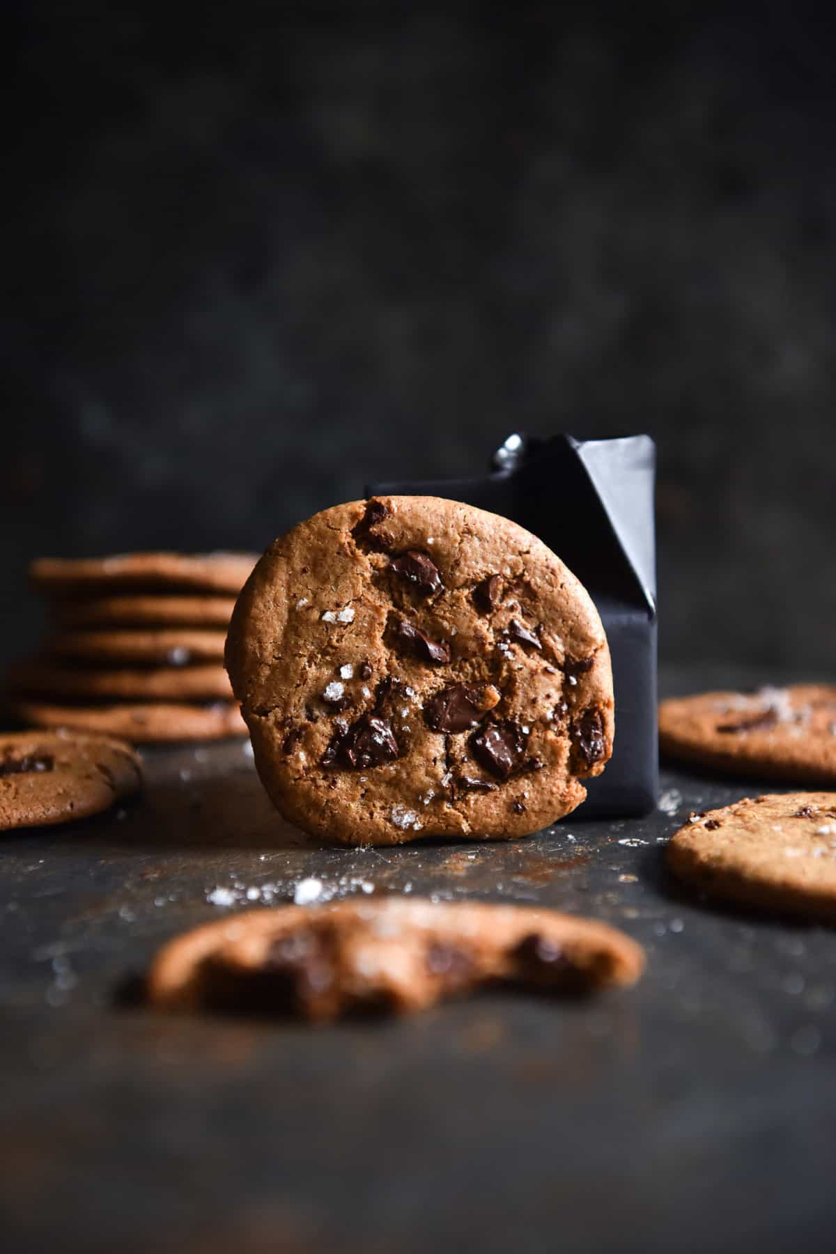 A moody side on image of an almond butter cookie against a dark coloured background. The central cookie faces the camera and is studded with chocolate chips. The remaining cookies lean up against the shelf around the central cookie.