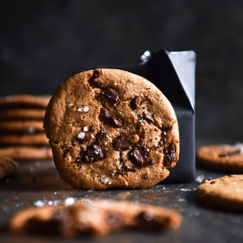 Vegan almond butter cookies studded with choc chips on a dark moody backdrop. One of the cookies is propped up against a black ceramic milk carton, showing the top of the cookie oozing with melted chocolate and topped with sea salt flakes.