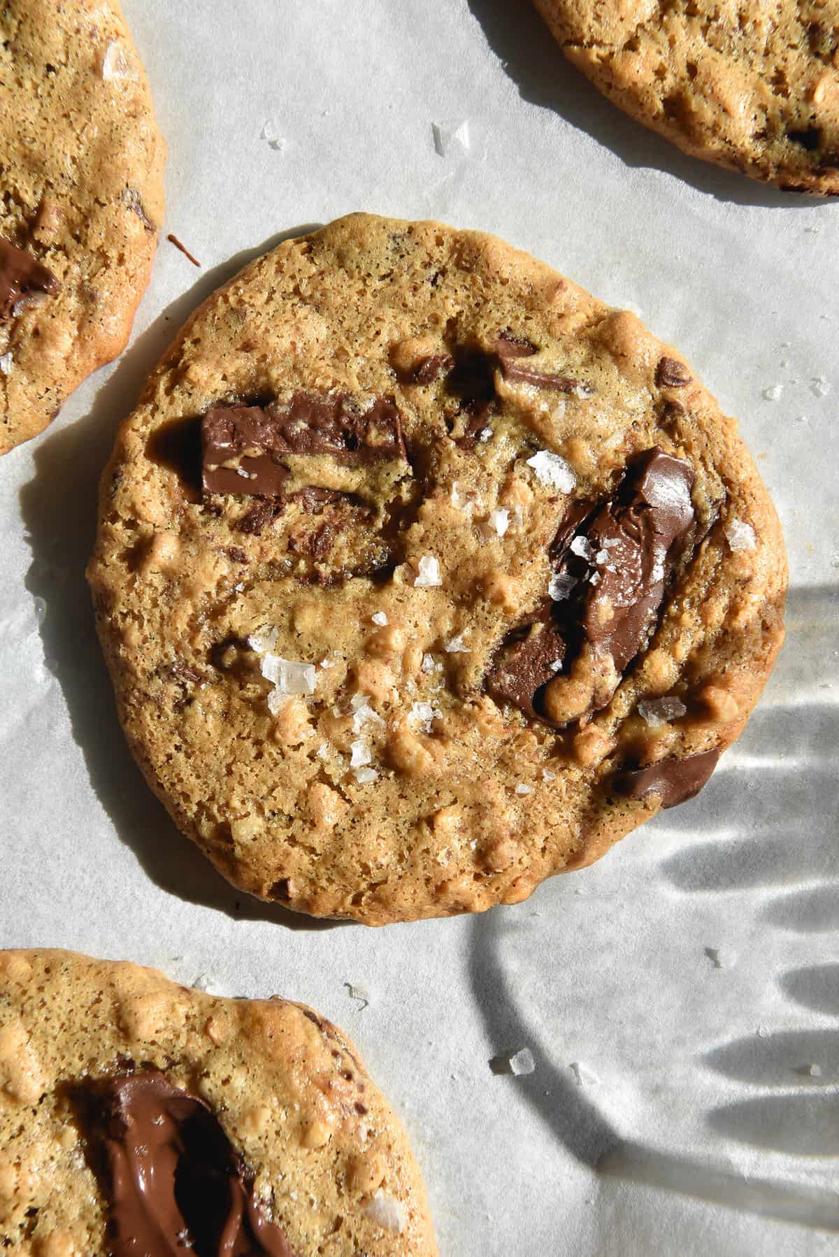 An aerial sunlit view of vegan peanut butter choc chip cookies on a white sheet of baking paper. 