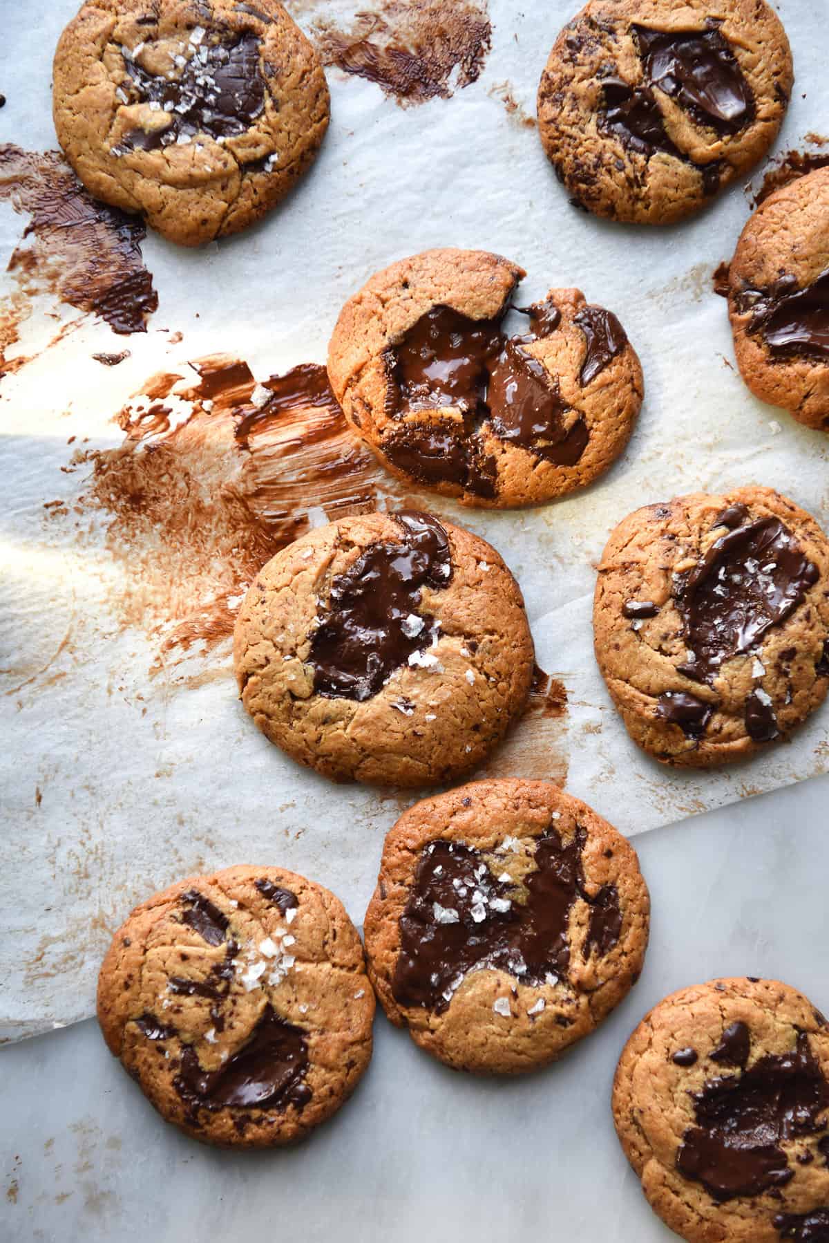 An aerial image of gluten free peanut butter cookies on baking paper atop a white marble table. The cookies are golden brown and studded wth melted chocolate.