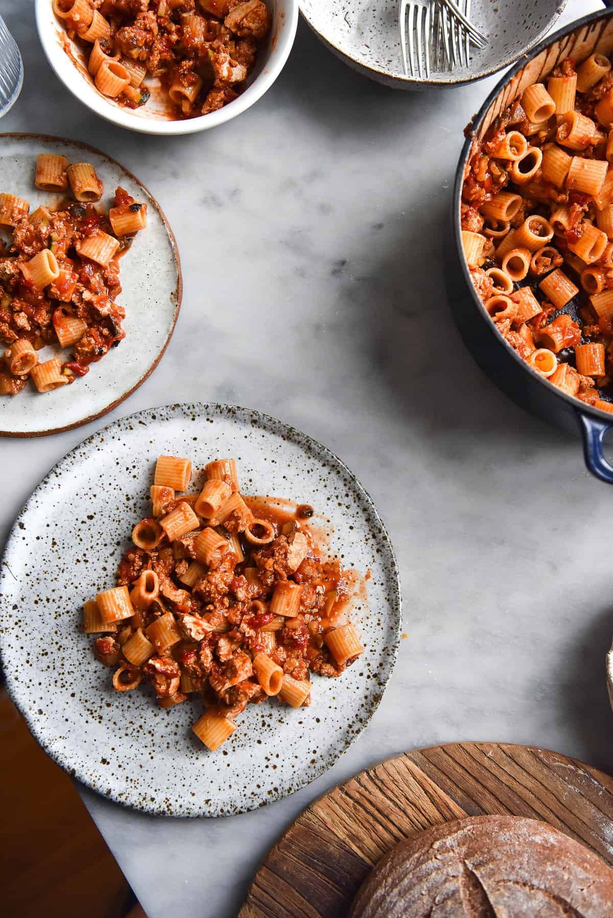 An aerial image of vegan bolognese on rigatoni pasta. The central plate is a white ceramic speckled plate that sits atop a white marble table. It is surrounded by other plates and a blue pot filled with more bolognese.