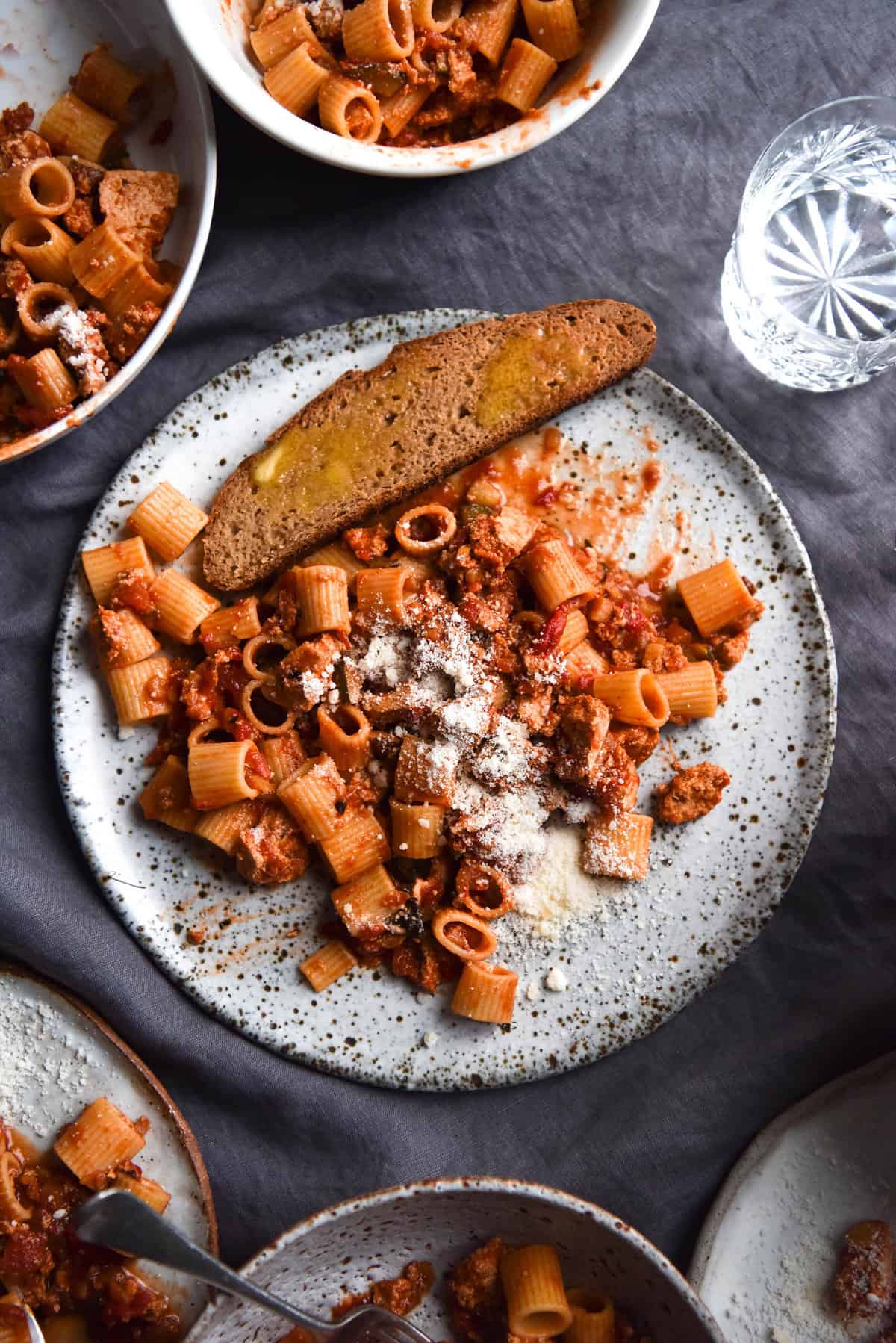 An aerial image of vegan bolognese on rigatoni pasta. The central plate of pasta sits atop a white speckled ceramic plate and has a sliced of buttered gluten free sourdough sitting at the top of the plate. The plate sits atop a purple linen tablecloth and is surrounded by extra bowls of bolognese.