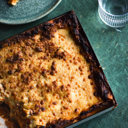 A FODMAP friendly vegan lasagne in a white rectangular baking dish on an olive green backdrop. A slice of the lasagne sits to the top left of the tray and a glass of water sits in the top right corner.