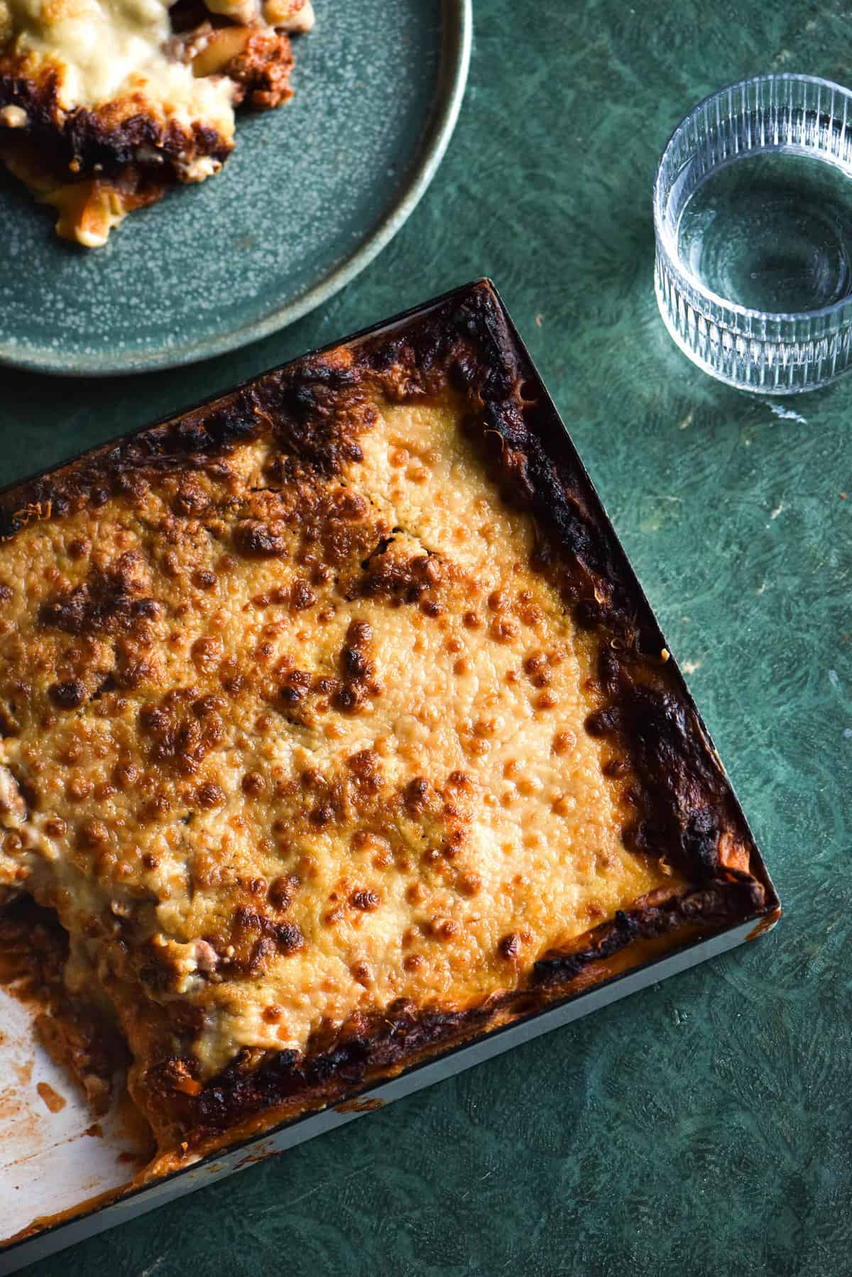 A FODMAP friendly vegan lasagne in a white rectangular baking dish on an olive green backdrop. A slice of the lasagne sits to the top left of the tray and a glass of water sits in the top right corner.