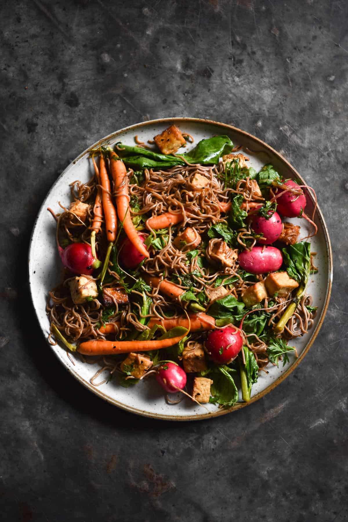An aerial image of sweet and sticky buckwheat noodles with tofu, radishes, carrots and Asian greens on a white ceramic plate atop a dark blue steel backdrop.