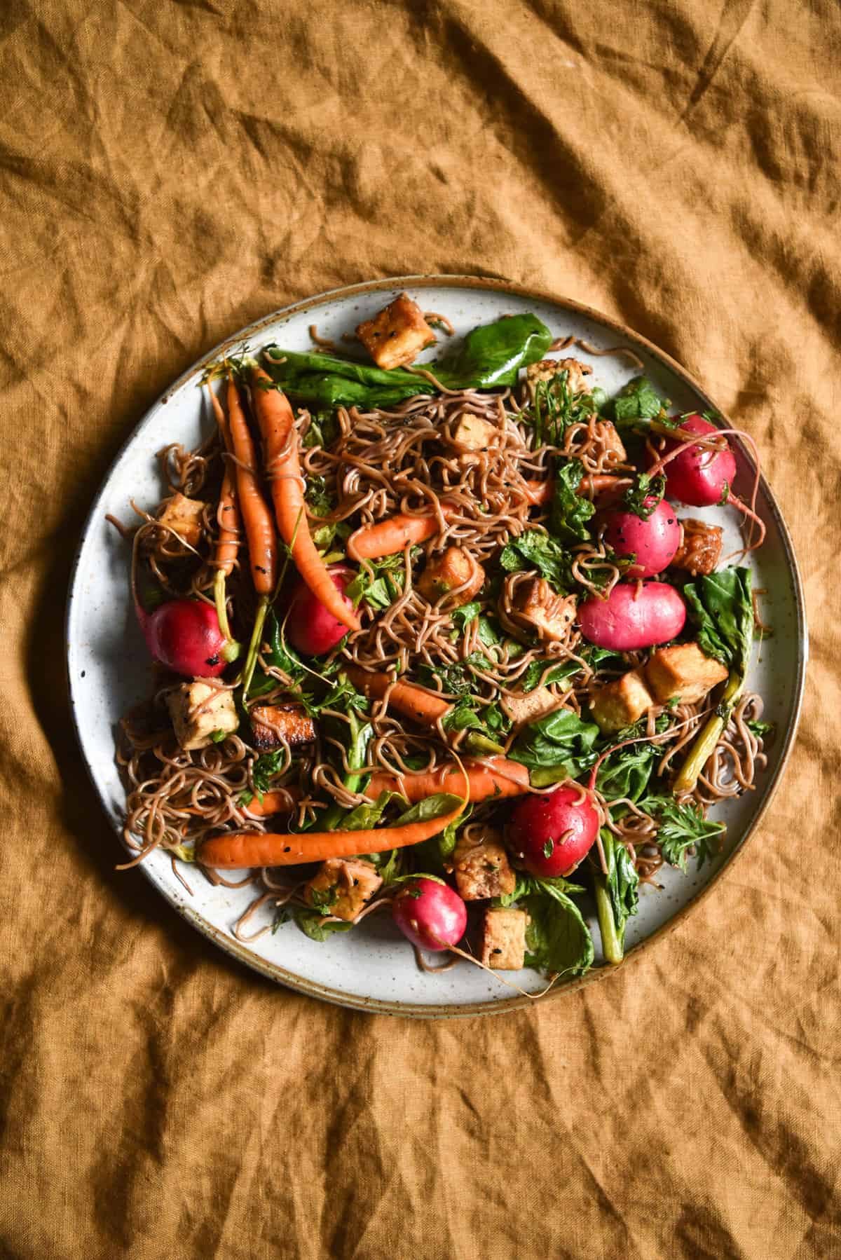 An aerial image of sweet and sticky buckwheat noodles with tofu, radishes, carrots and Asian greens on a white ceramic plate atop a turmeric coloured linen tablecloth. 