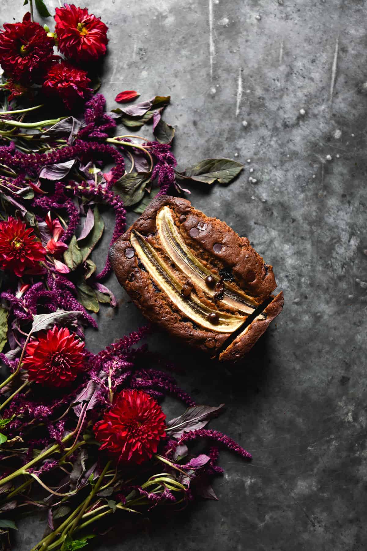 An aerial image of a loaf of gluten free banana bread on a dark blue steel backdrop surrounded by red and purple flowers.