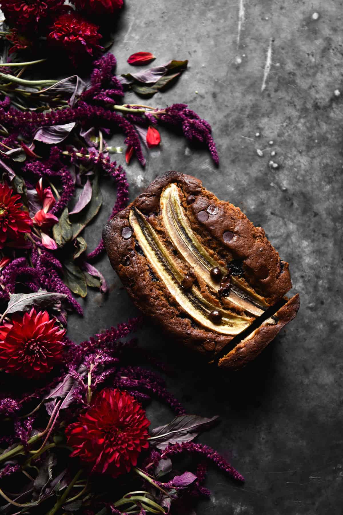 An aerial image of a loaf of gluten free banana bread on a dark blue steel backdrop surrounded by red and purple flowers.