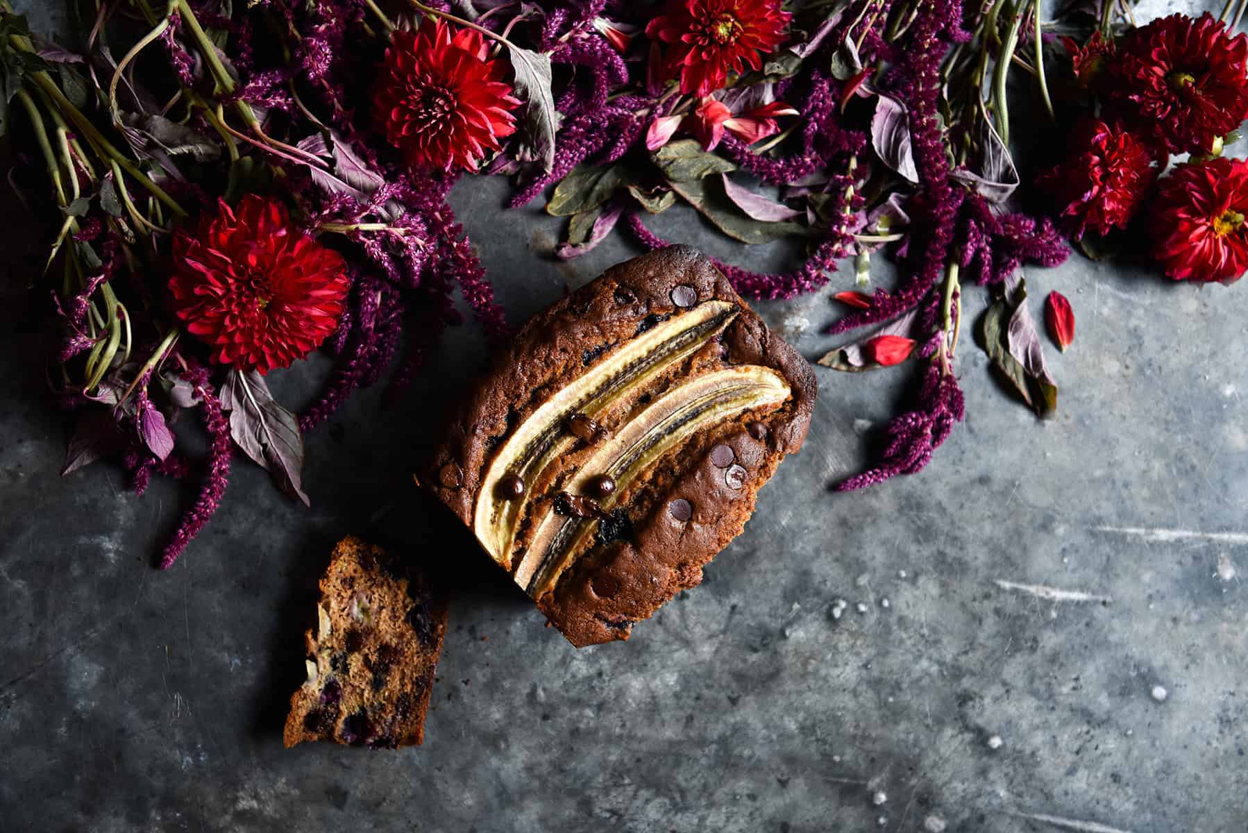 An aerial image of a loaf of gluten free banana bread on a dark blue steel backdrop surrounded by red and purple flowers.