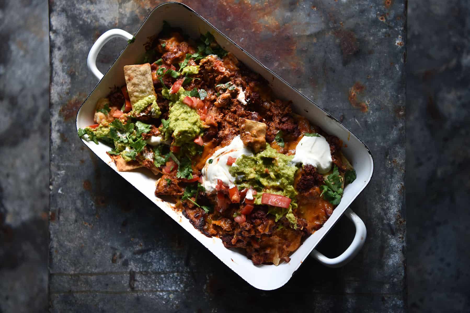 An aerial image of a tray of nachos on a dark blue steel backdrop. The nachos are topped with vegetarian mince, queso, sour cream, guacamole and a tomato coriander salsa.