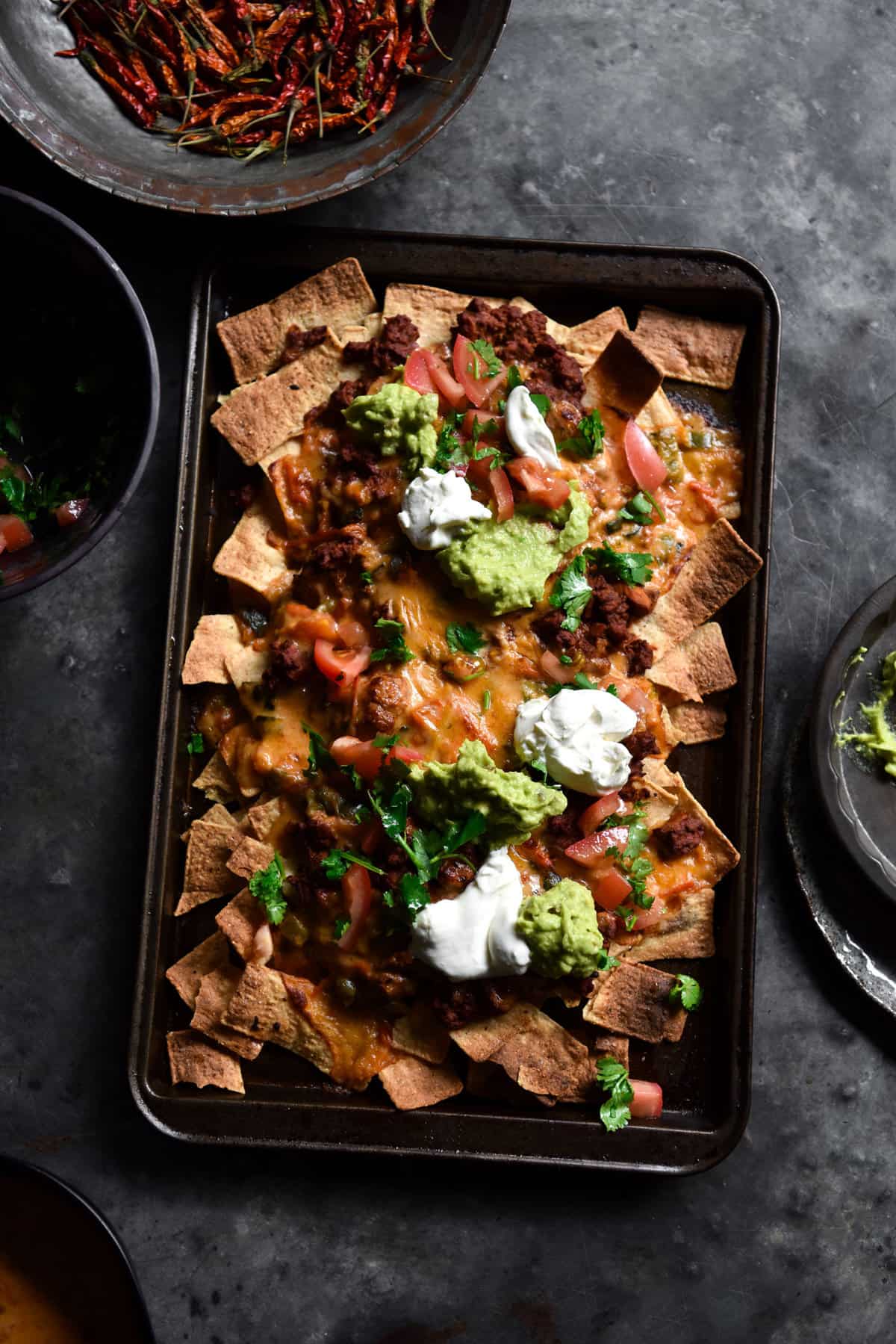An aerial image of a tray of nachos on a dark blue steel backdrop. The nachos are topped with vegetarian mince, queso, sour cream, guacamole and a tomato coriander salsa.