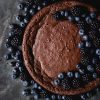 A flourless chocolate cake topped with a crescent moon shape design of blueberries and blackberries. The cake sits atop a dark blue mottled steel backdrop and is surrounded by extra berries.