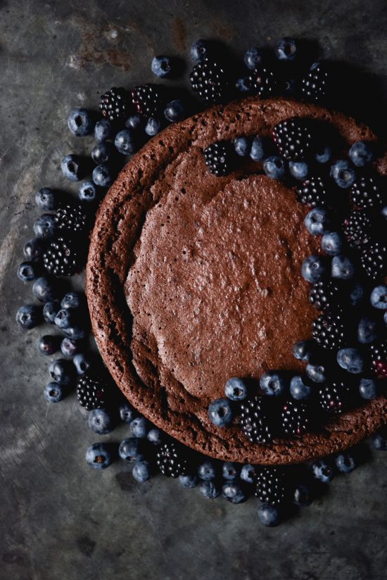 A flourless chocolate cake topped with a crescent moon shape design of blueberries and blackberries. The cake sits atop a dark blue mottled steel backdrop and is surrounded by extra berries.
