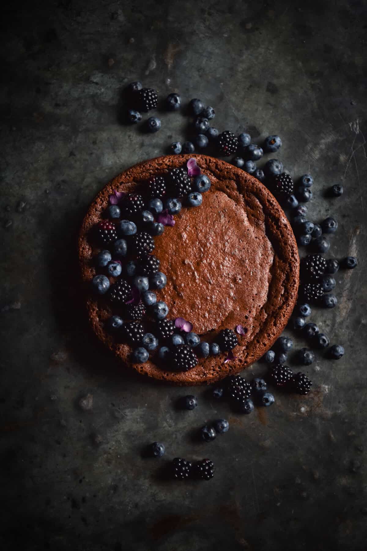 An aerial image of a flourless, nut free chocolate cake atop a dark steel backdrop. The cake has been decorated with blueberries, blackberries and purple flowers in a half moon shape on the left side of the cake.