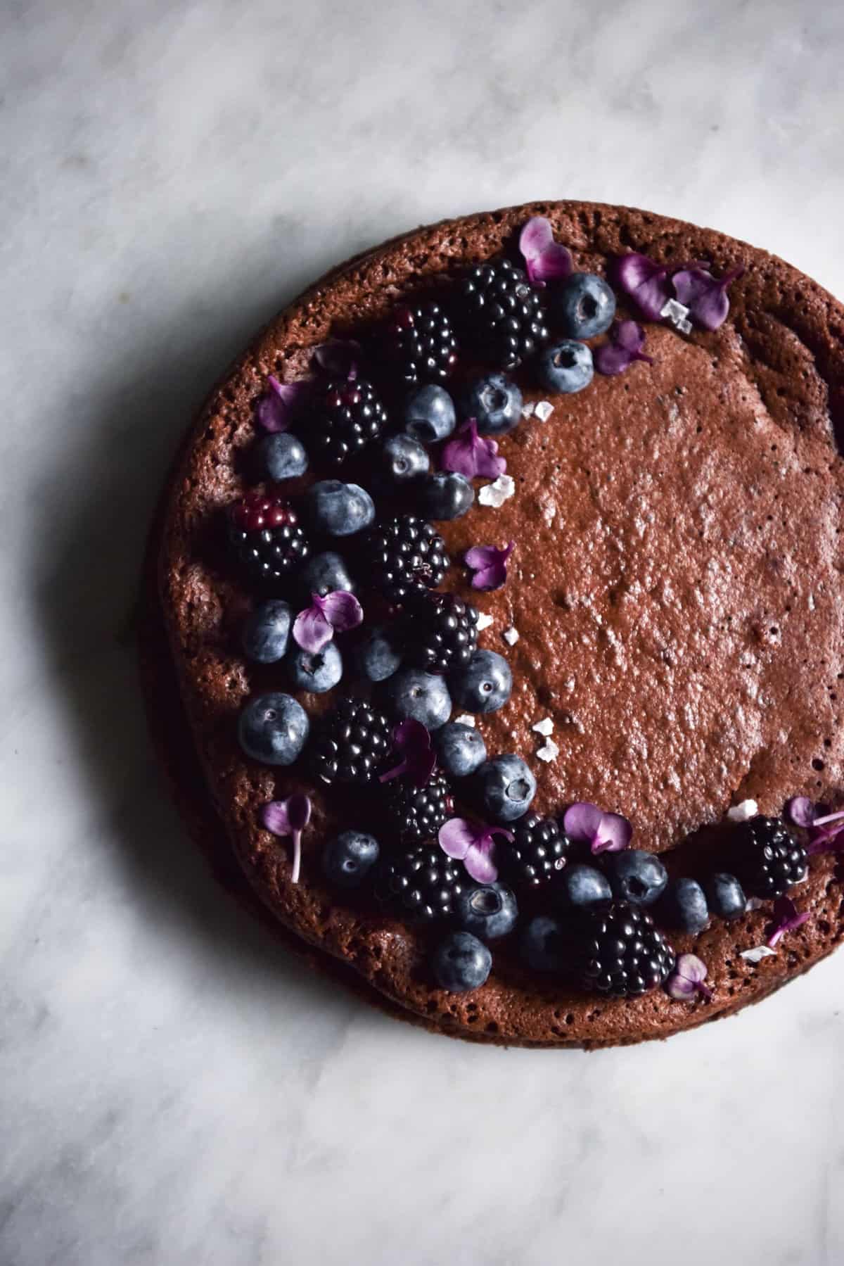 An aerial image of a flourless chocolate cake atop a white marble table. The cake is decorated with a crescent moon shape design of blueberries, blackberries and purple edible flowers.
