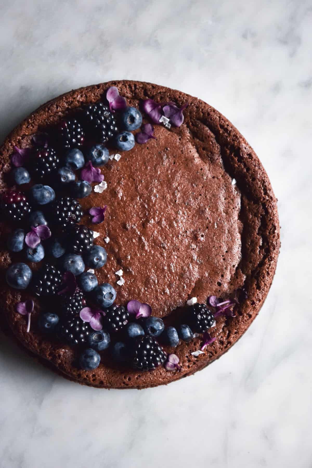 An aerial image of a flourless, nut free chocolate cake atop a white marble table. The cake has been decorated with blueberries, blackberries and purple flowers in a half moon shape on the left side of the cake.
