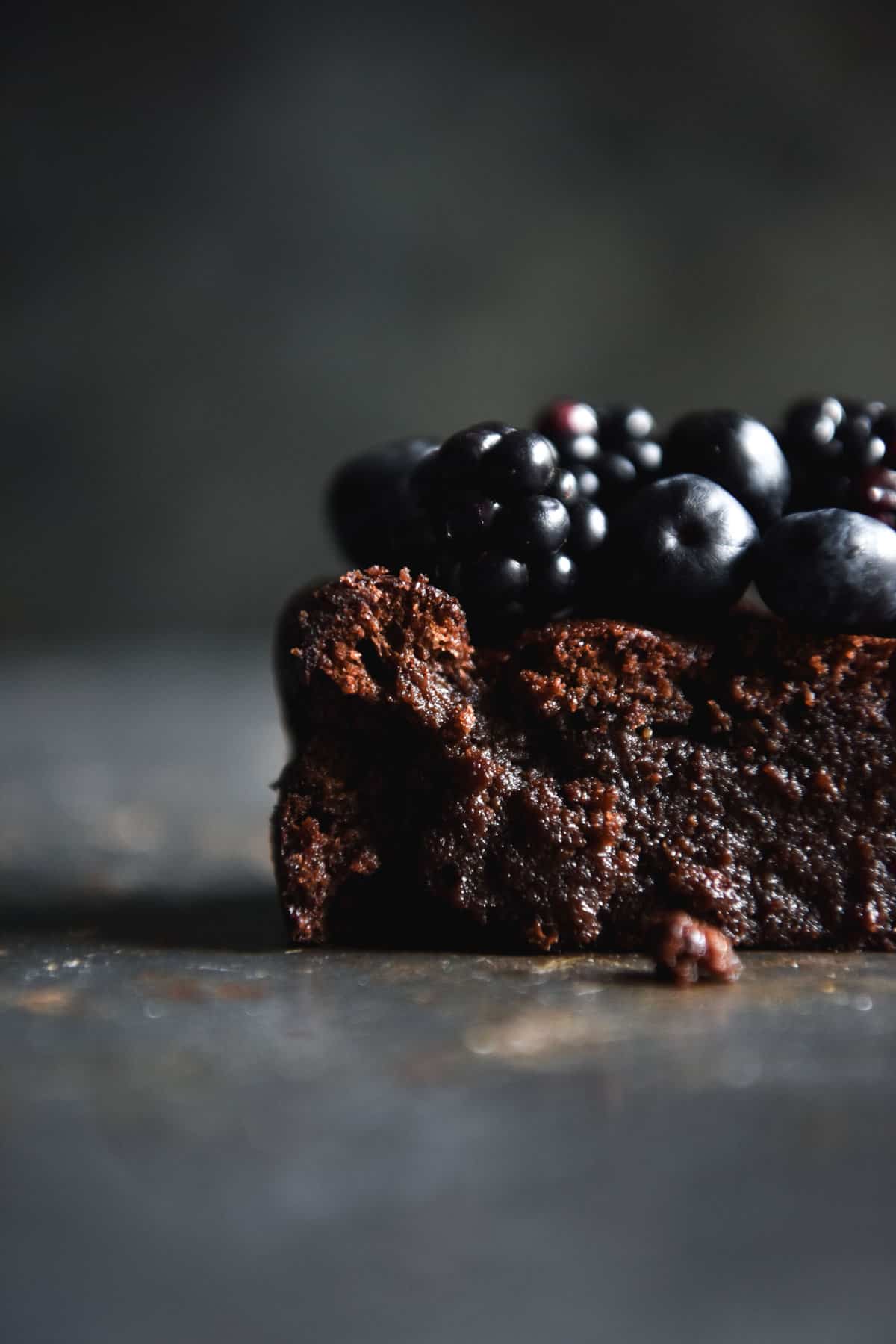 A side on close up of the fudgy insides of a flourless nut free chocolate cake. The piece of cake sits on a blue steel backdrop and is topped with fresh berries. 