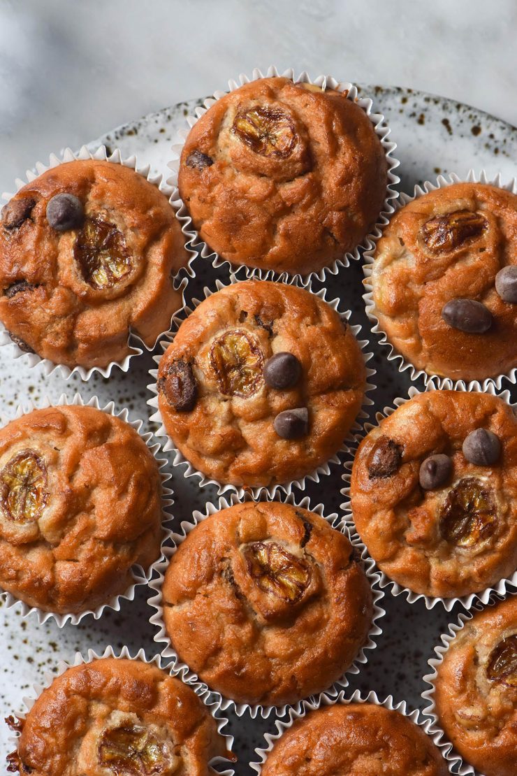 An aerial image of gluten free banana muffins on a white speckled ceramic plate atop a white marble table.