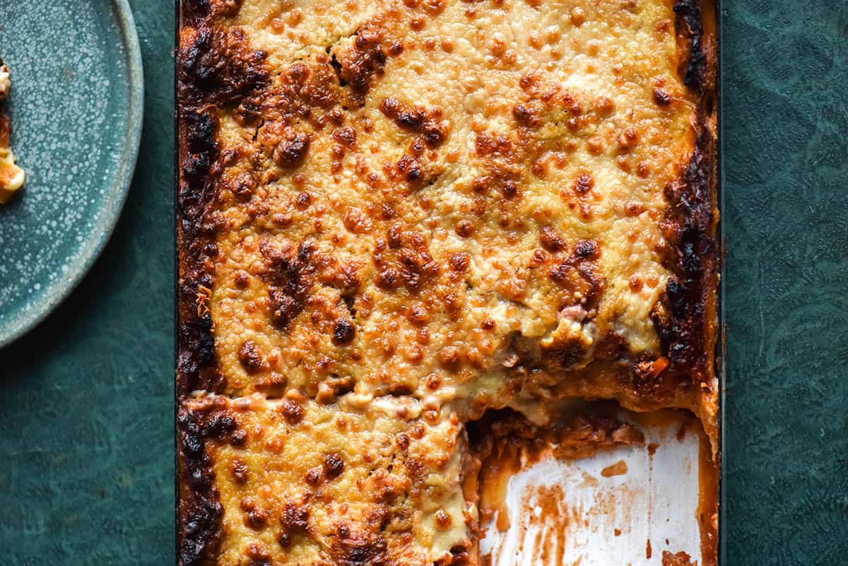 An aerial view of a gluten free vegan lasagne on a dark green backdrop. A piece of the lasagne has been removed from the bottom right side of the dish and served on a green ceramic plate to the top left of the image